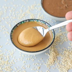 A spoon scoops creamy tahini from a small bowl, surrounded by scattered sesame seeds on a light surface. Another bowl with tahini is blurred in the background.