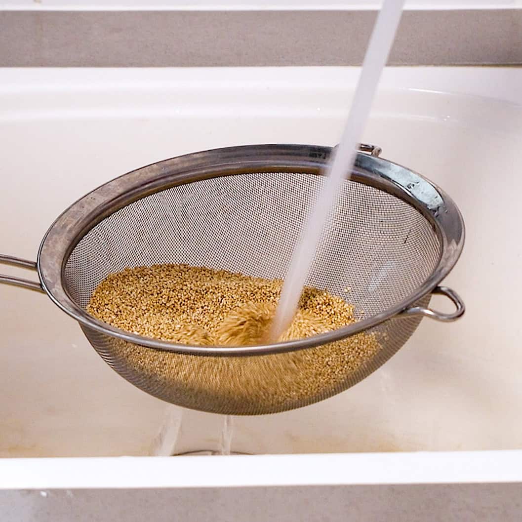 A metal strainer filled with quinoa is placed over a white sink, while water is being poured onto the quinoa to rinse it.