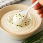 A hand holds a spoonful of creamy white sour cream dip garnished with chopped chives above a bowl filled with the same dip. A few fresh chives are placed on the table nearby.