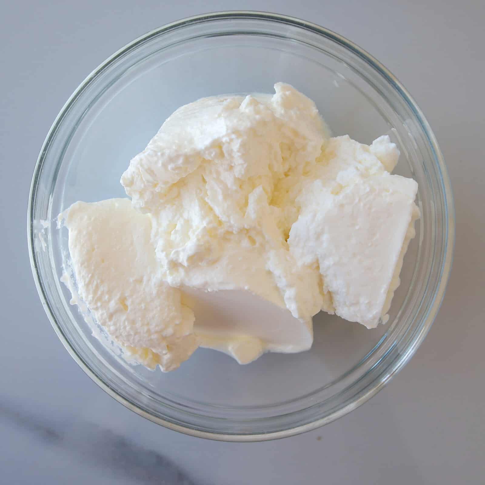 A clear glass bowl filled with several scoops of thick, white, creamy sour cream, on a light gray surface.