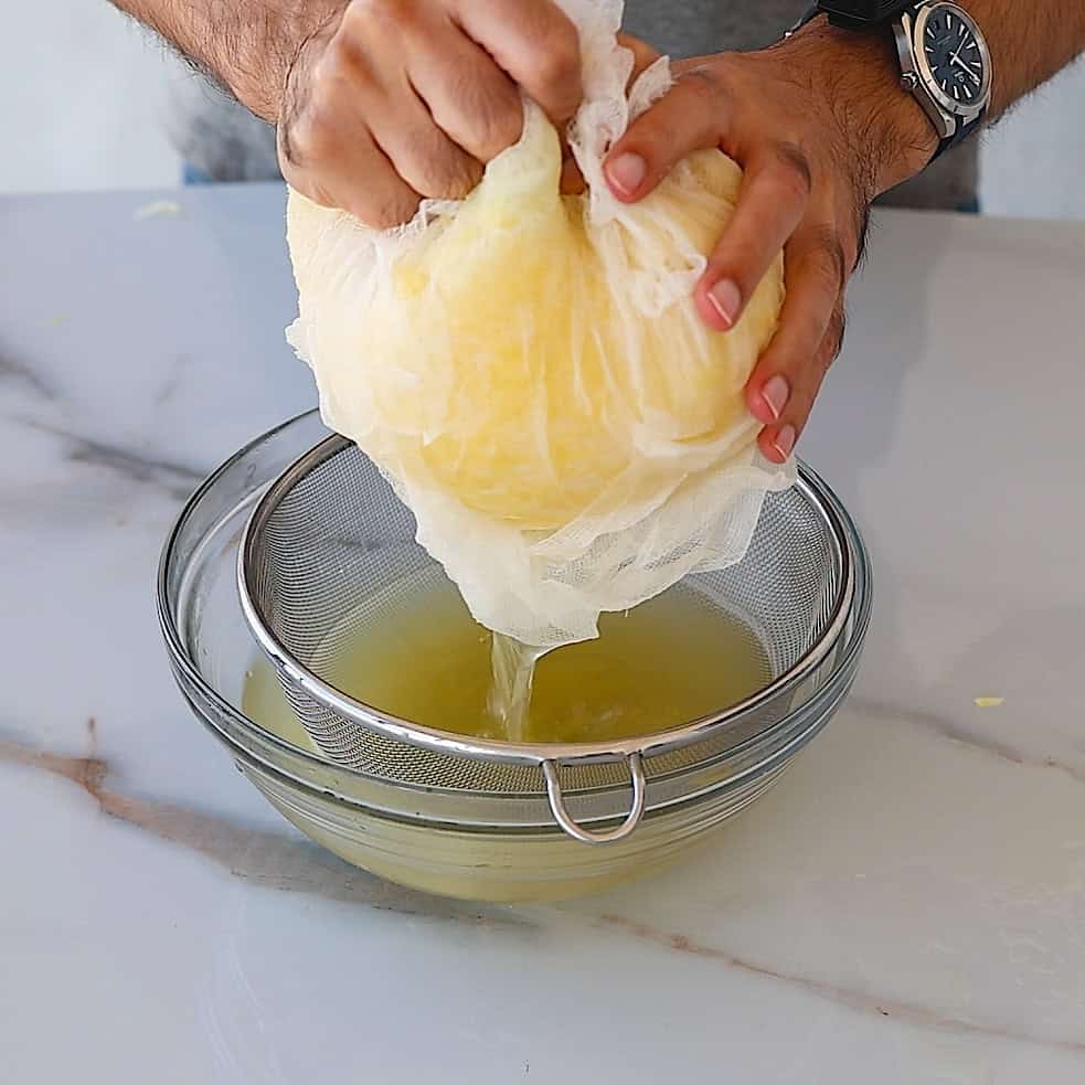 A person squeezes the liquids from grated potatoes in a cloth over a sieve and glass bowl, straining out liquid on a white countertop.