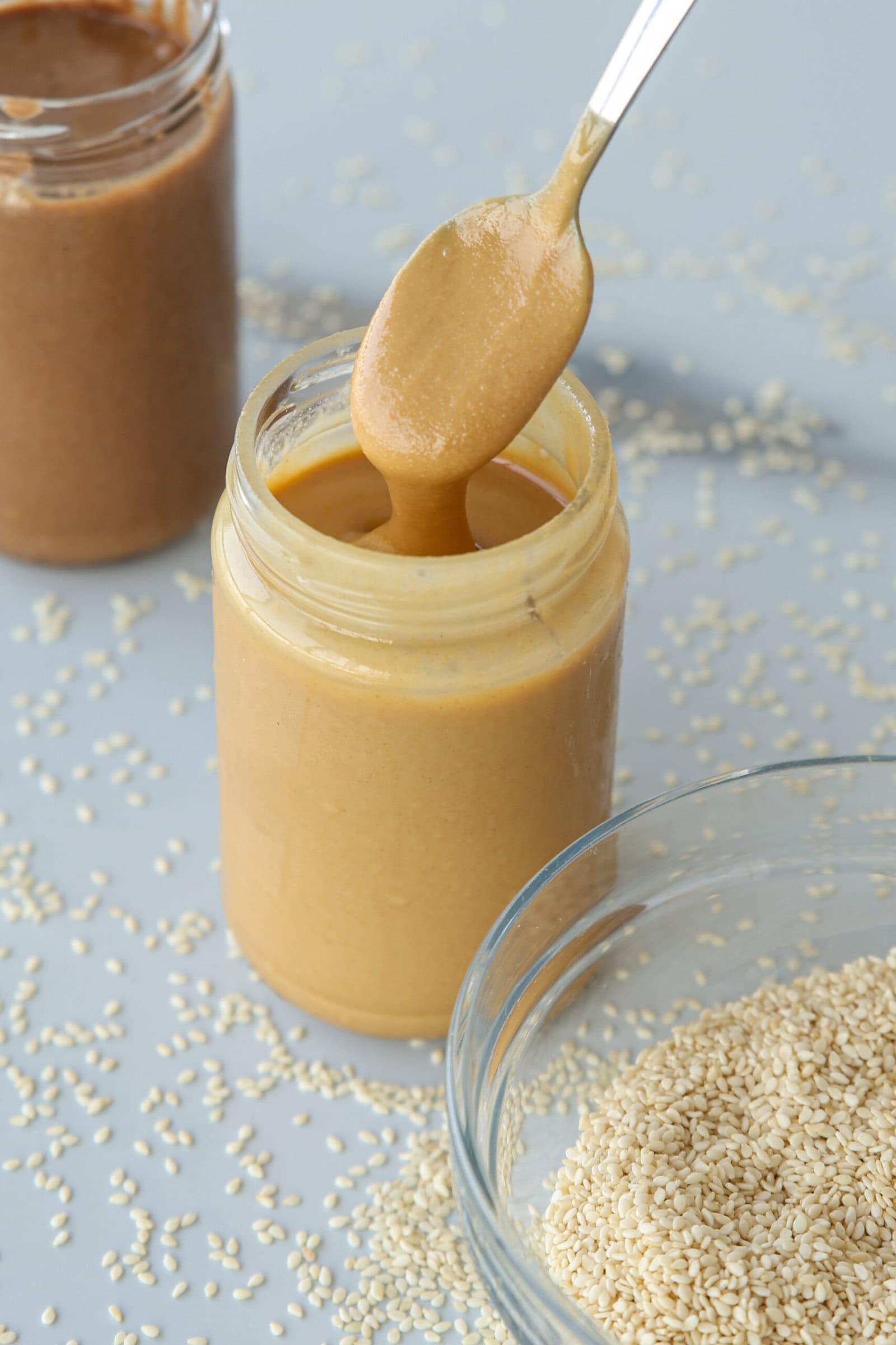 A spoon with creamy tahini is held above an open jar, with sesame seeds scattered on the surface and a bowl of sesame seeds nearby. Another jar is visible in the background.