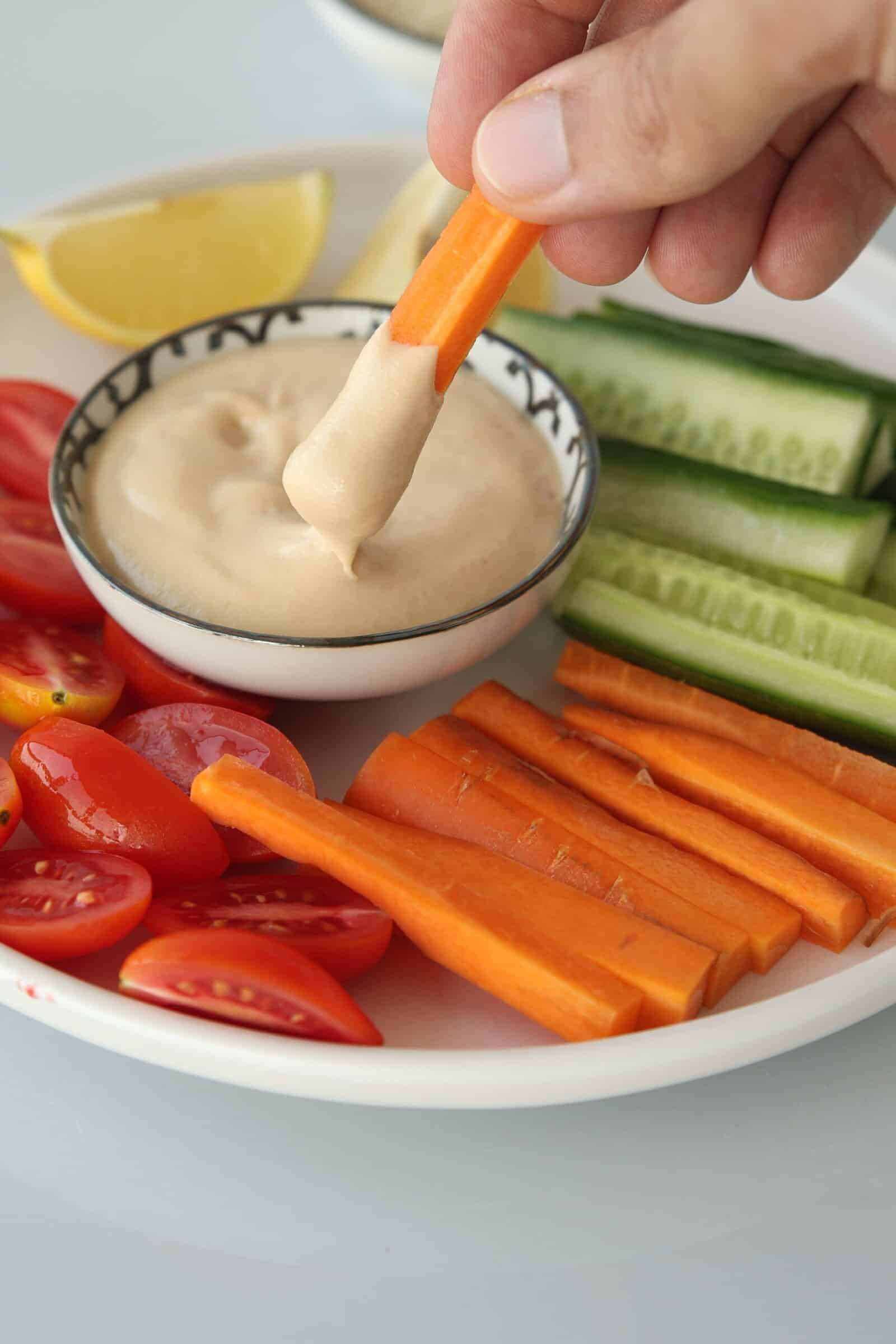 A hand dips a carrot stick into a bowl of creamy tahini sauce, surrounded by sliced cucumbers, tomatoes, and more carrot sticks on a white plate. A lemon wedge is also visible on the plate.