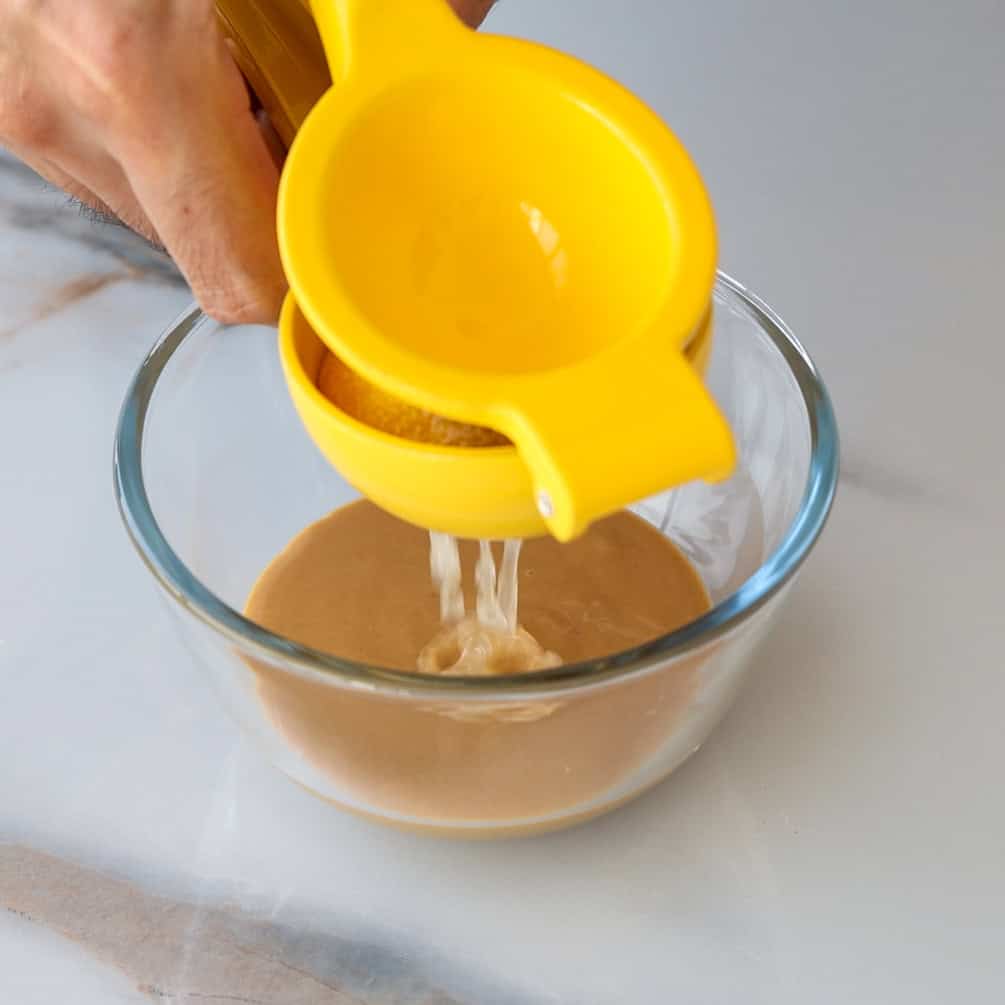 A hand squeezes a yellow citrus juicer, pouring juice into a glass bowl containing a light brown liquid on a white surface.