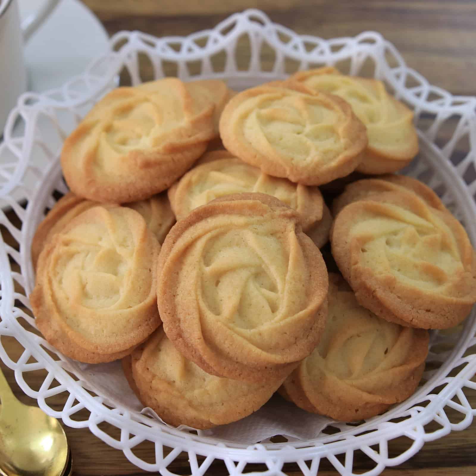 A white basket filled with round, swirled melt-in-your-mouth butter cookies sits on a wooden table next to a gold spoon.