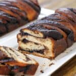 A close-up of sliced chocolate babka on a white rectangular plate, showing its swirled chocolate filling and golden-brown crust. Another loaf is visible in the background, slightly out of focus.