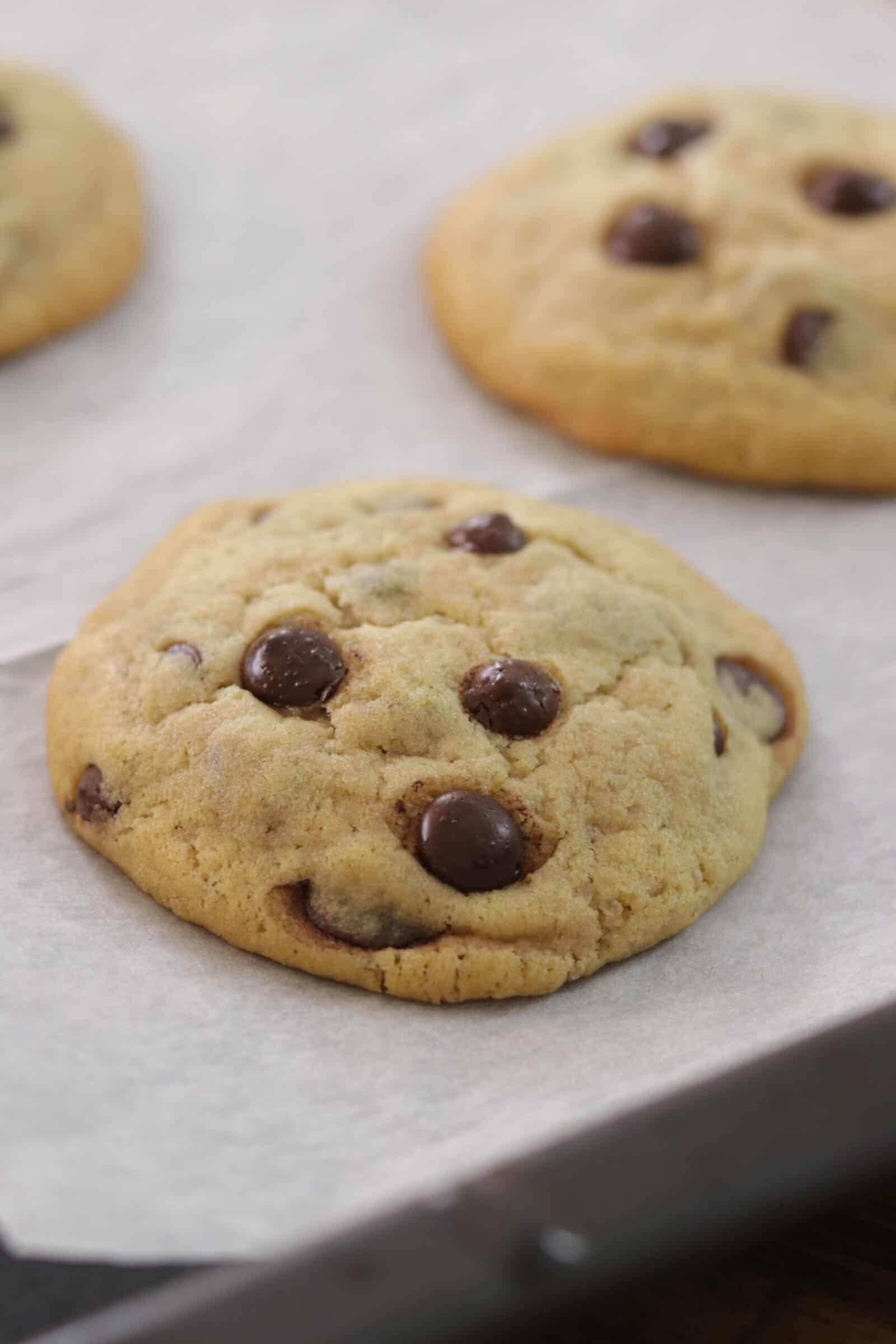 A close-up of a freshly baked chocolate chip cookie on parchment paper, with other cookies blurred in the background.