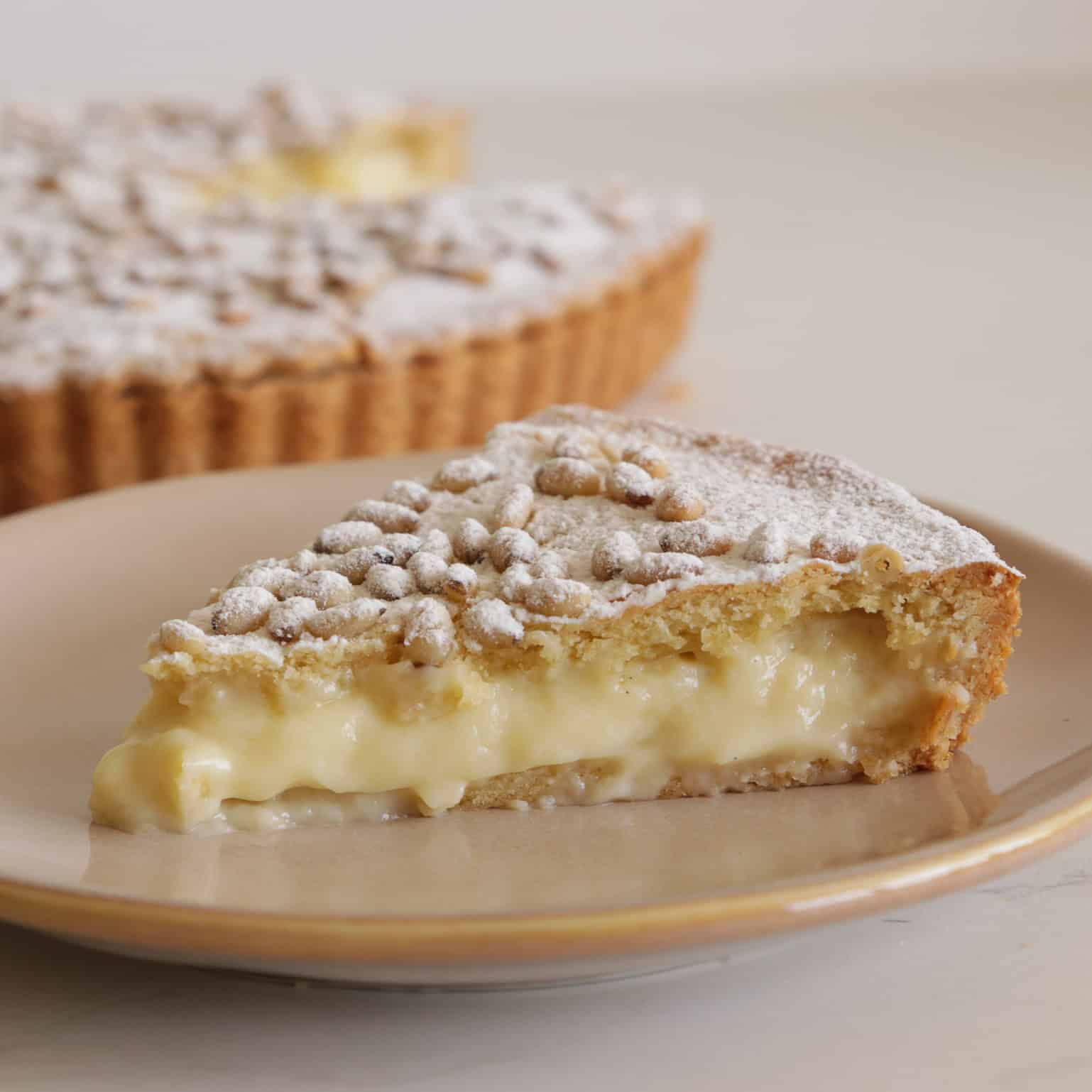 A slice of creamy custard pie with a golden (torta della nonna), crumbly crust, dusted with powdered sugar, sits on a beige plate. The rest of the pie is visible in the background.