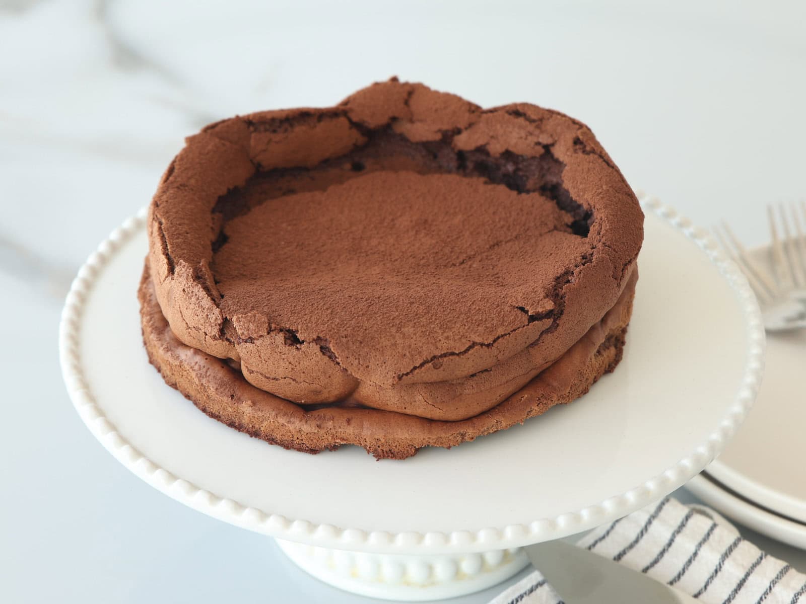 A cracked, flourless chocolate cake dusted with cocoa powder sits on a white cake stand. A striped napkin and forks are visible beside the stand, with a blurred white background.