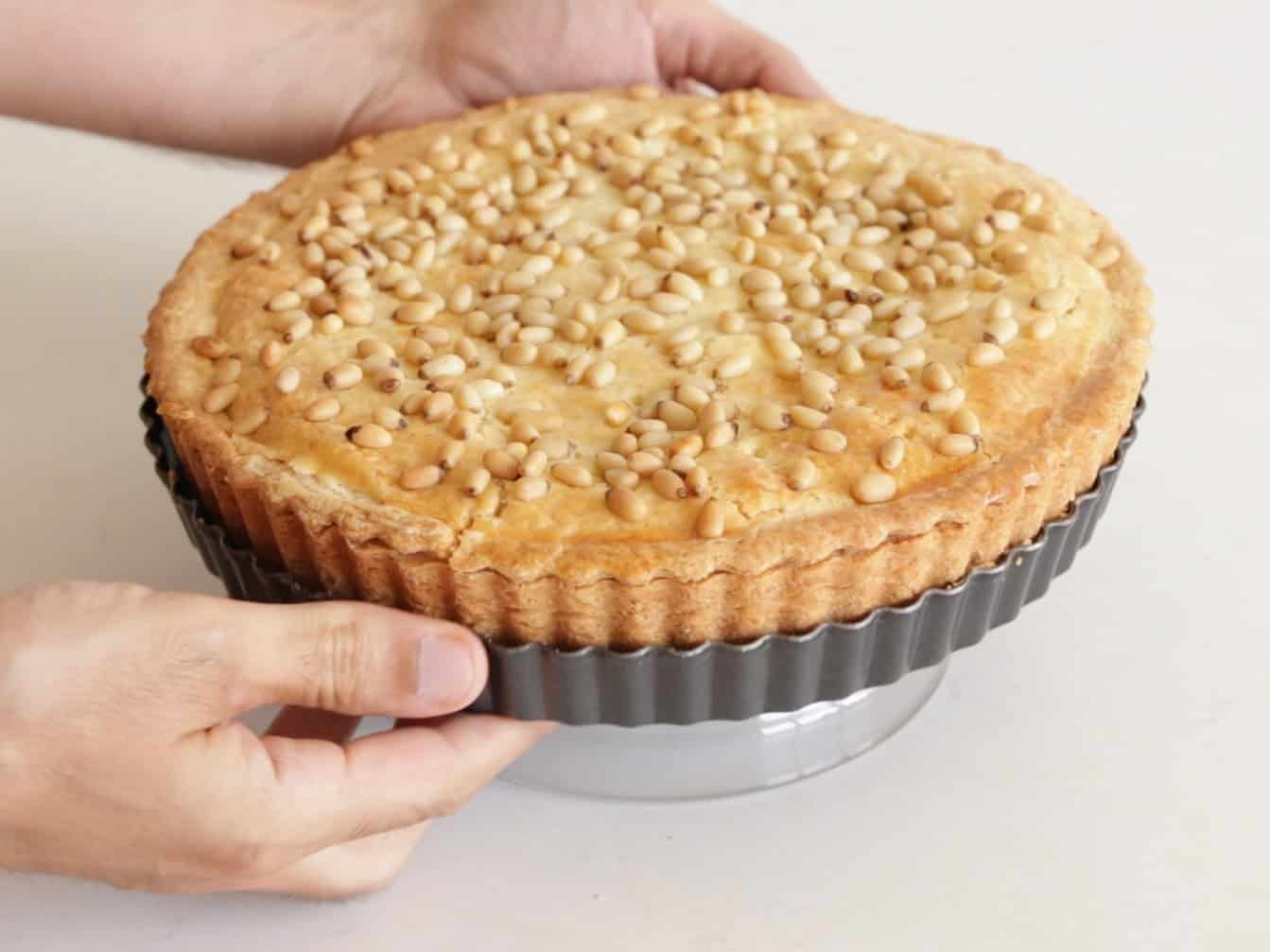 A person lifts a freshly baked tart covered with pine nuts out of a metal tart pan, onto a glass stand, against a light background.