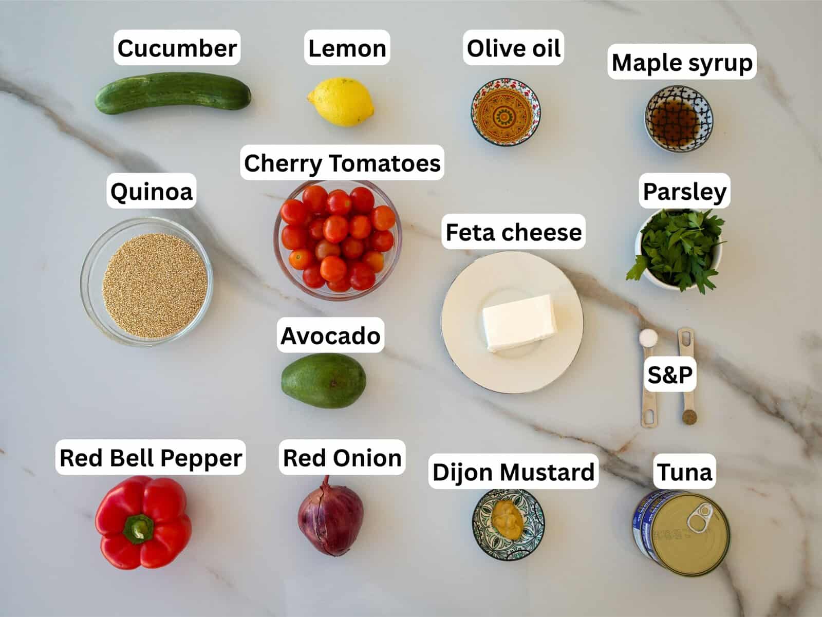 A top-down view of labeled ingredients on a white surface, including cucumber, lemon, olive oil, maple syrup, quinoa, cherry tomatoes, feta cheese, parsley, avocado, red bell pepper, red onion, Dijon mustard, salt & pepper, and tuna.