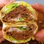 A close-up of hands holding a wrap cut in half, showing layers of doner kebab meat, lettuce, onions, and sauce inside a tortilla.