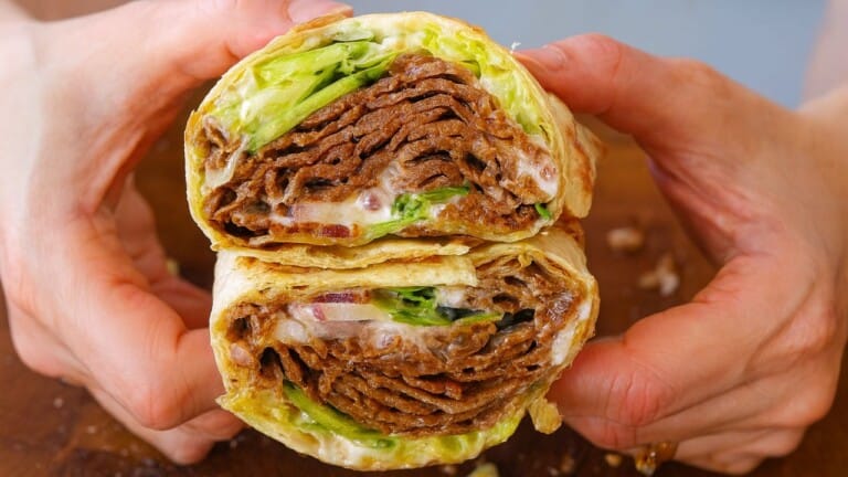 A close-up of hands holding a wrap cut in half, showing layers of doner kebab meat, lettuce, onions, and sauce inside a tortilla.