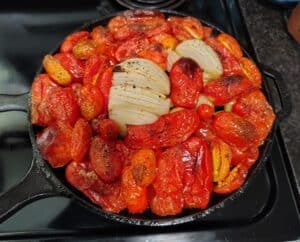 A cast iron skillet filled with roasted tomatoes and a halved onion, both seasoned with pepper and salt. The vegetables are slightly charred and arranged neatly on a stovetop.