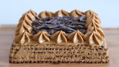 Easy Chocotorta Recipe | Classic Argentinian No-Bake Cake A square layered cake with coffee-colored frosting, topped with piped dollops and chocolate shavings, sits on a wooden surface. The cake has a textured, airy appearance with visible layers.