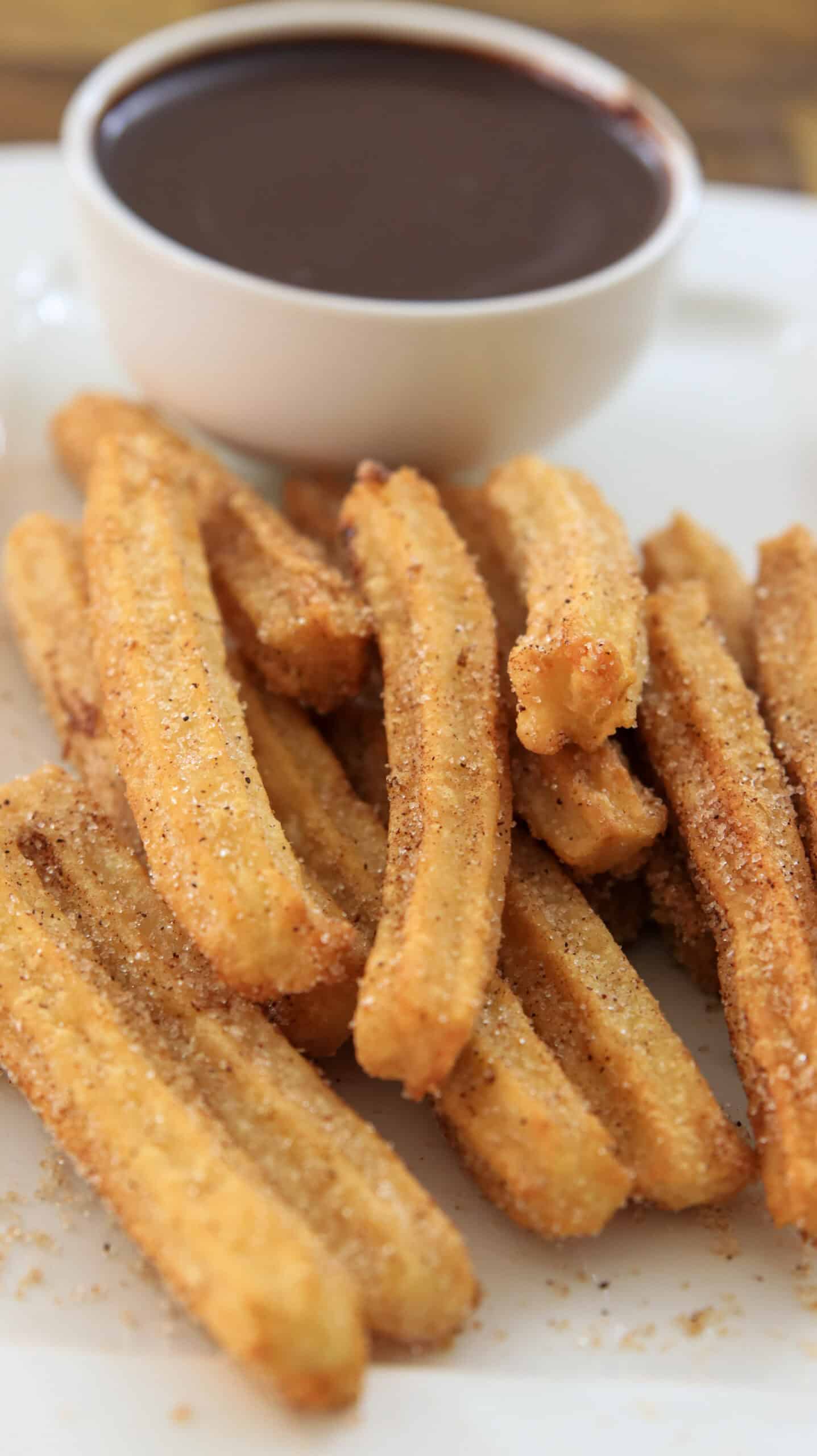 A plate of golden, sugar-coated churros is served with a small white cup filled with rich chocolate dipping sauce.