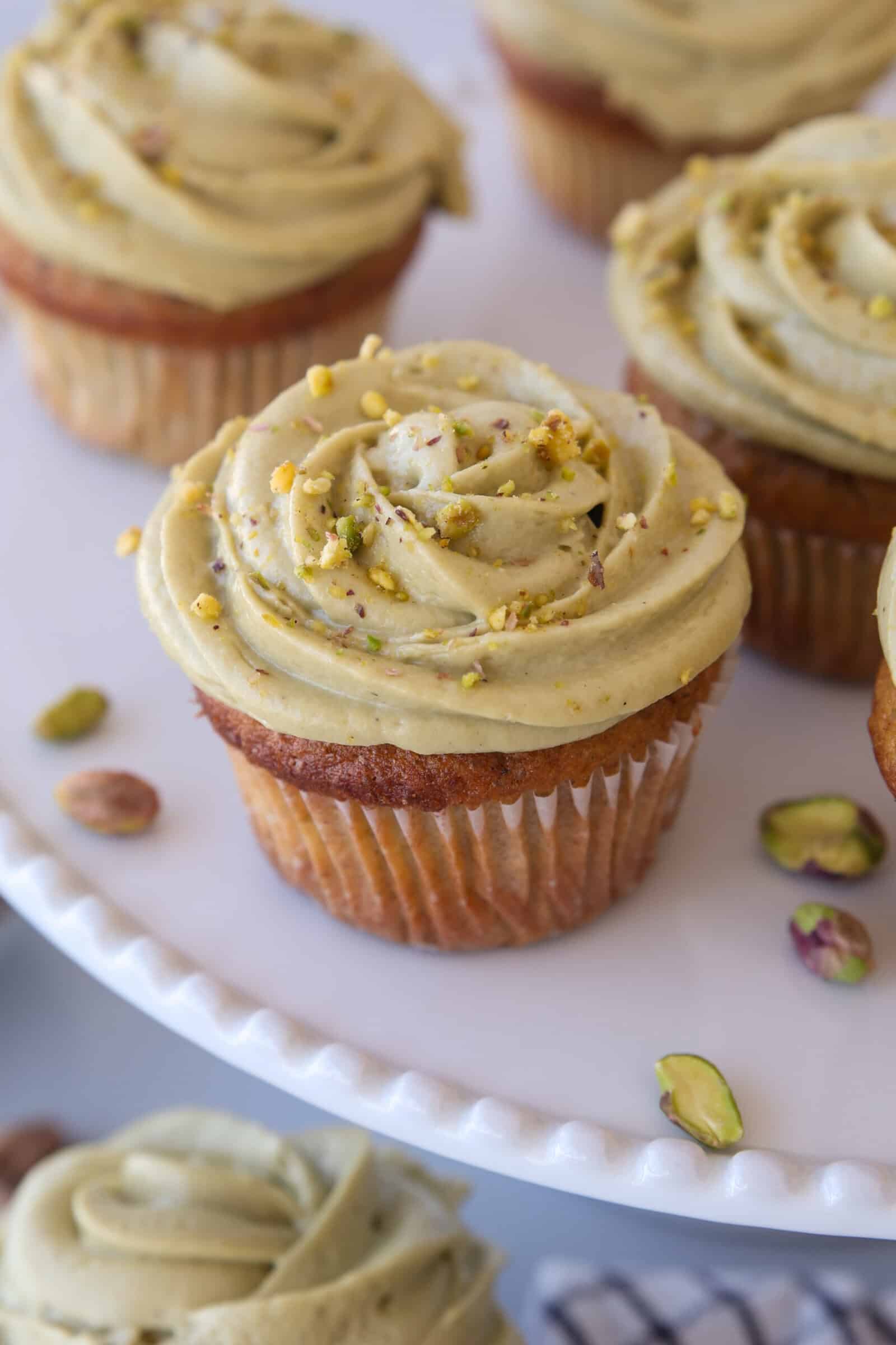 A close-up of cupcakes on a white plate, topped with light green pistachio frosting and sprinkled with crushed pistachios. Some whole pistachios are scattered nearby.