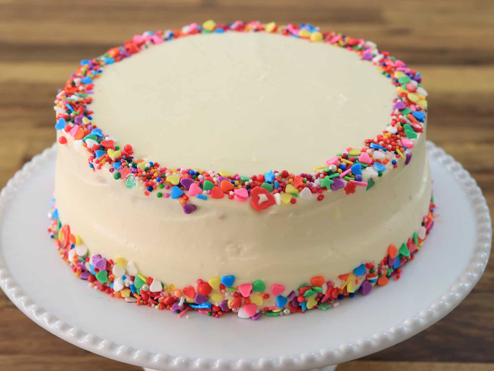 A round cake with smooth white frosting, decorated with colorful sprinkles along the top and bottom edges, sits on a white cake stand with a wooden surface in the background.