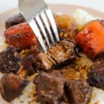 A close-up of a fork cutting into a piece of tender beef stew on a bed of white rice, with visible chunks of carrots and mushrooms covered in rich brown gravy.