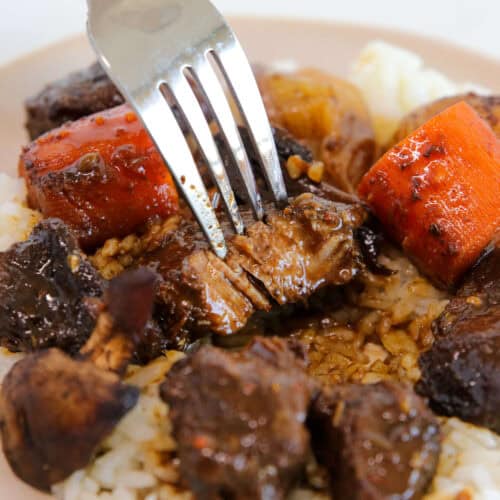 A close-up of a fork cutting into a piece of tender beef stew on a bed of white rice, with visible chunks of carrots and mushrooms covered in rich brown gravy.