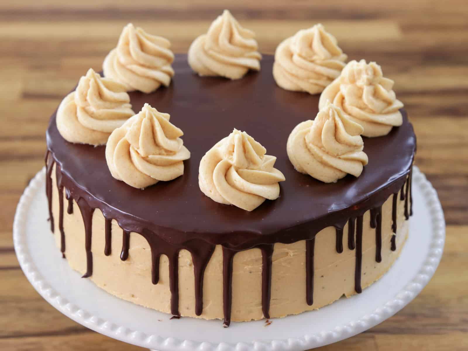 A round cake with smooth light brown frosting and chocolate ganache dripping down the sides, topped with eight evenly spaced swirls of piped frosting, displayed on a white cake stand.