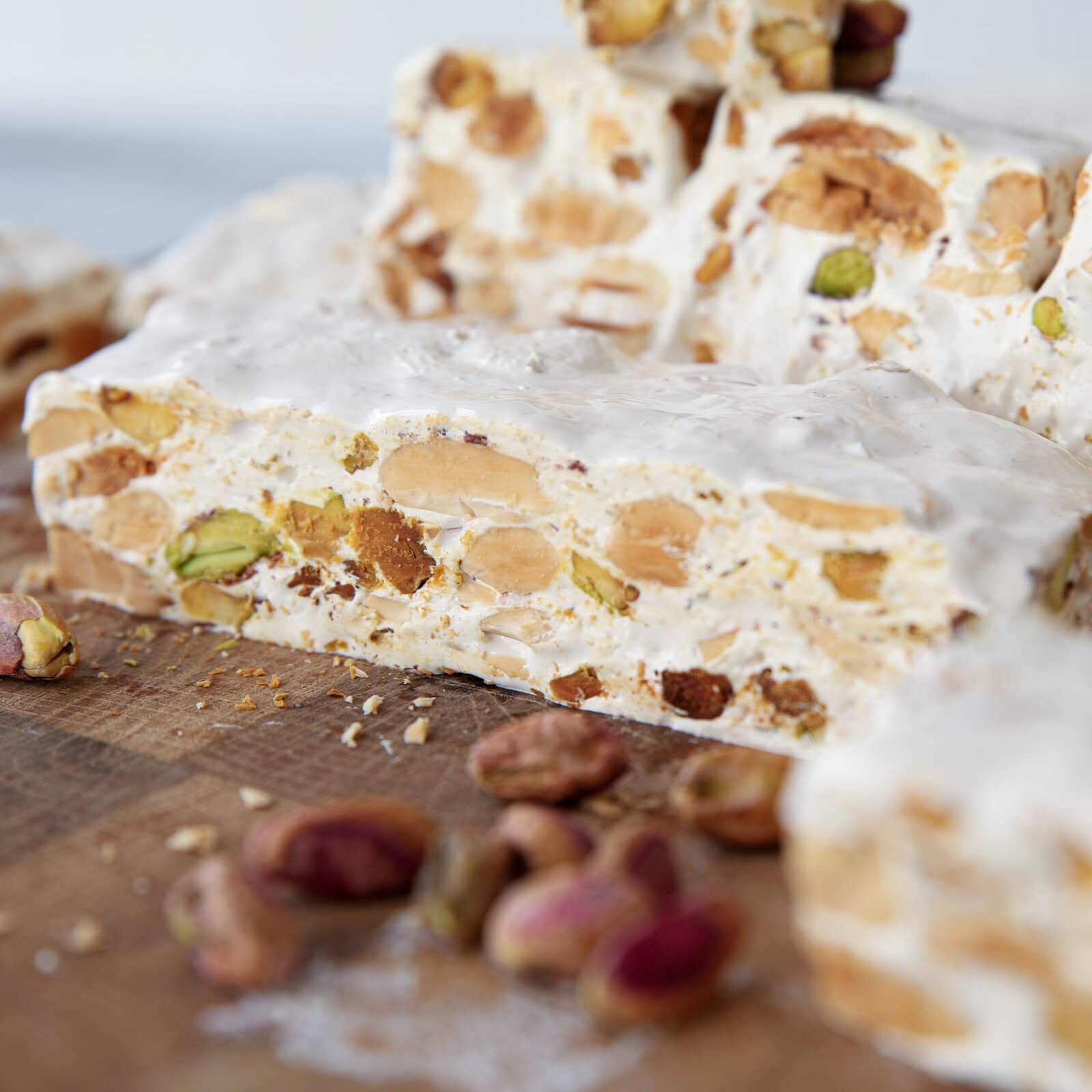 Close-up of nougat slices with visible nuts, including pistachios and almonds, on a wooden surface. Some whole pistachios are scattered in the foreground.