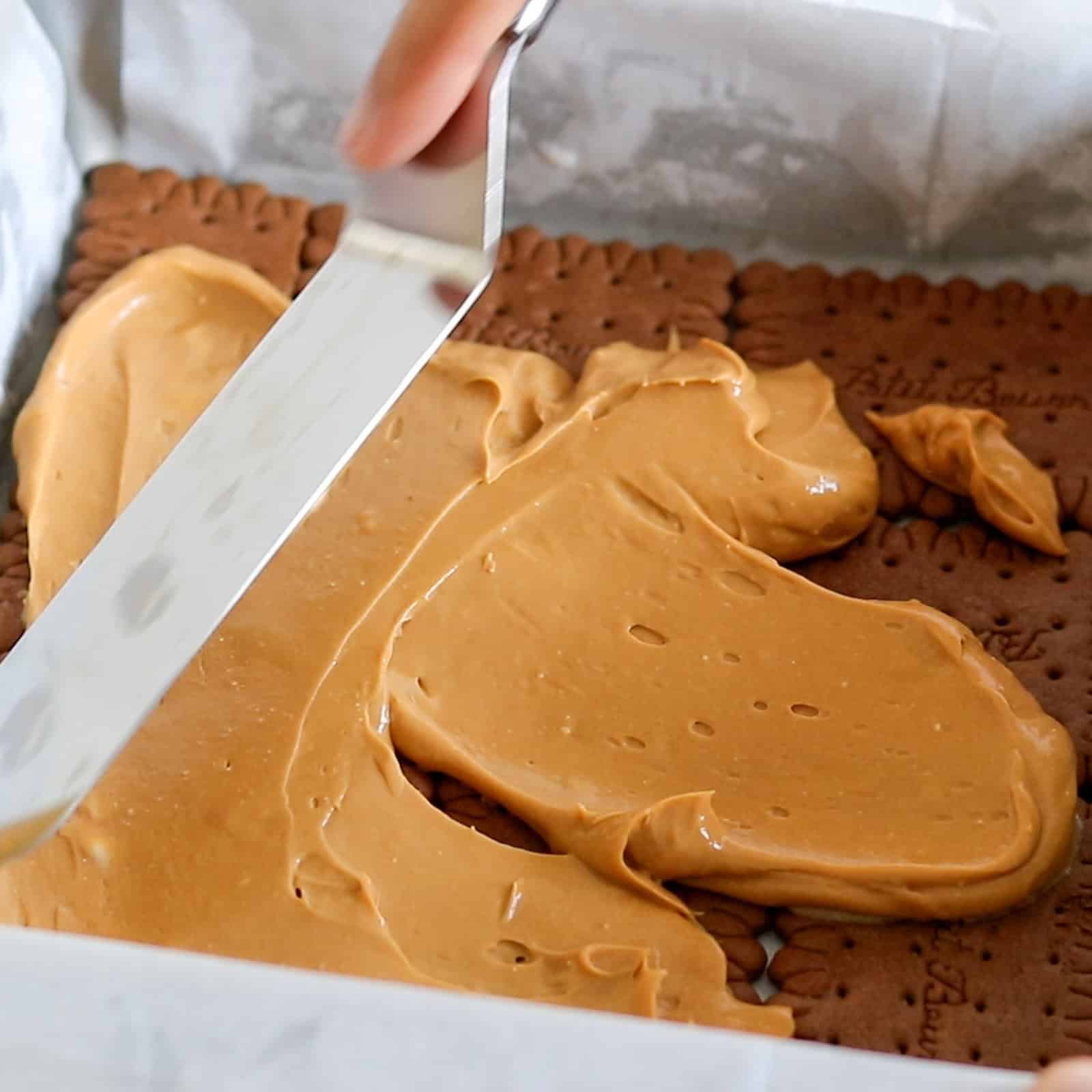 A hand uses a spatula to spread creamy caramel or dulce de leche over a layer of rectangular biscuits in a baking tray lined with parchment paper.