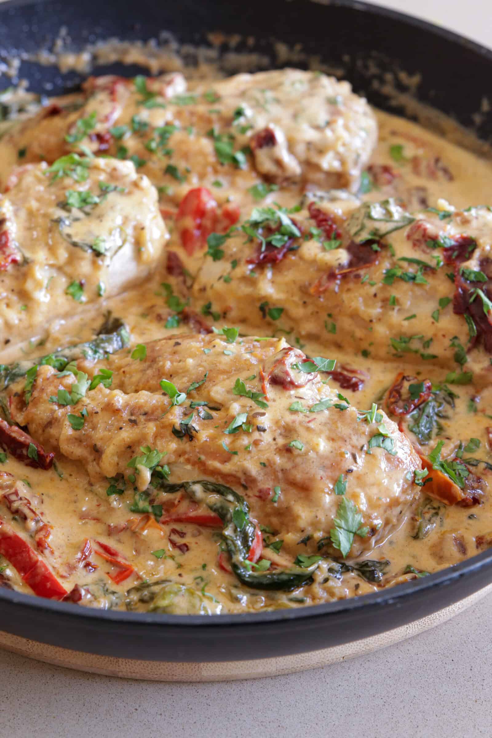 Four pieces of Tuscan-style chicken breast cooked in a creamy sauce with spinach, sun-dried tomatoes, herbs, and garnished with chopped parsley in a black skillet.