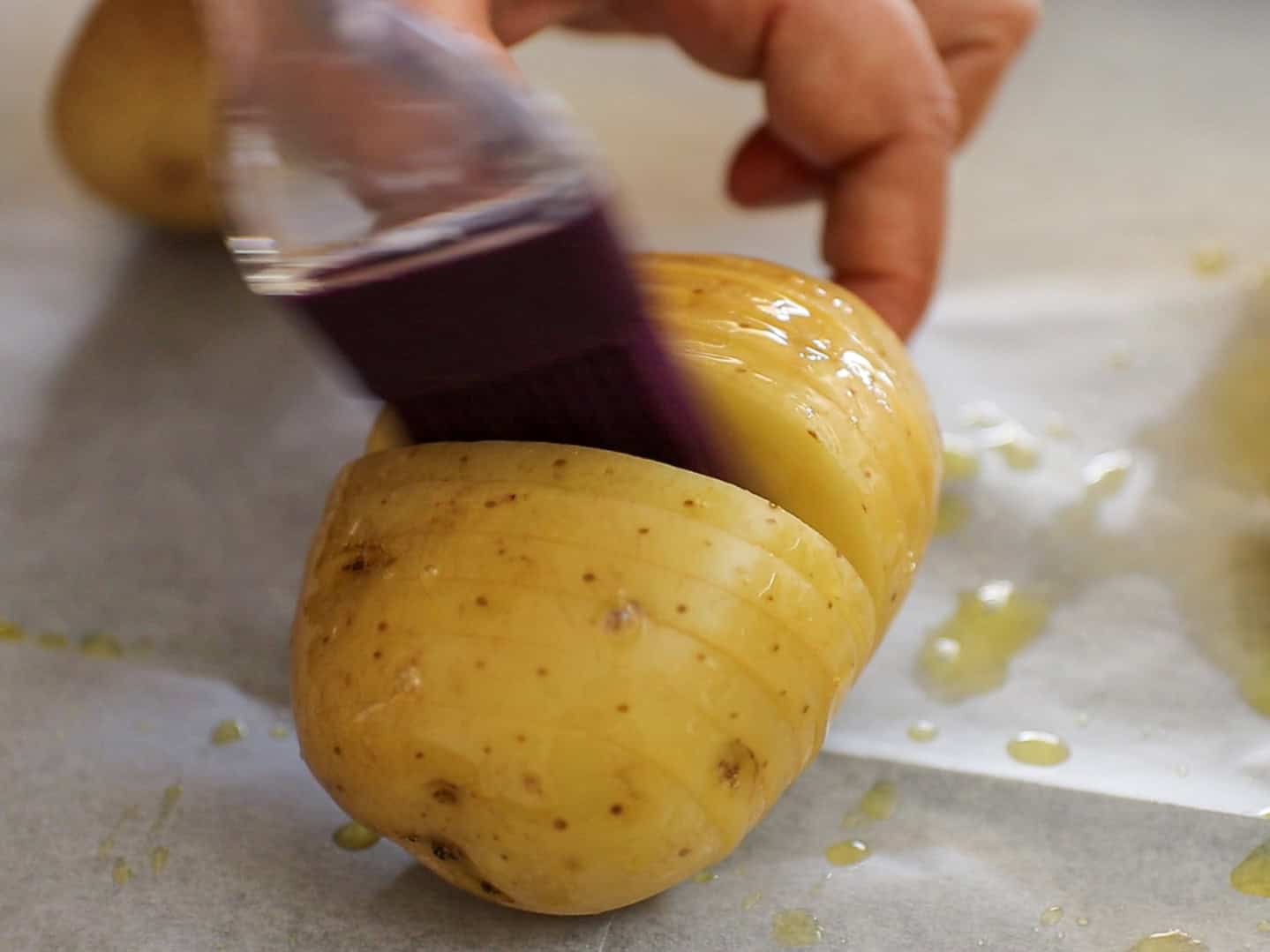 A hand uses a purple brush to apply oil to a sliced potato on parchment paper, preparing it for roasting.