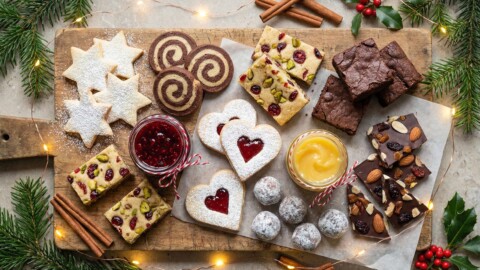 49 Easy Homemade Food Gifts for Christmas (Edible and Beautiful) A wooden board with assorted holiday cookies, brownies, jam, and lemon curd, surrounded by pine branches, cinnamon sticks, holly, and fairy lights.