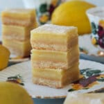 Three stacked lemon bars with a crumbly base and yellow filling are displayed on a floral-patterned plate, surrounded by fresh lemons and a teacup with a similar flower design.