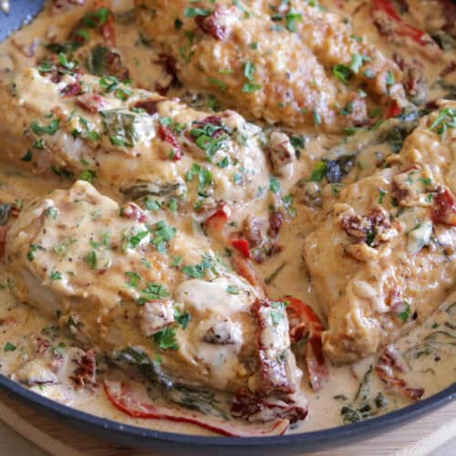 Chicken breasts simmering in a creamy sauce with spinach, sun-dried tomatoes, and herbs, garnished with chopped parsley in a skillet.