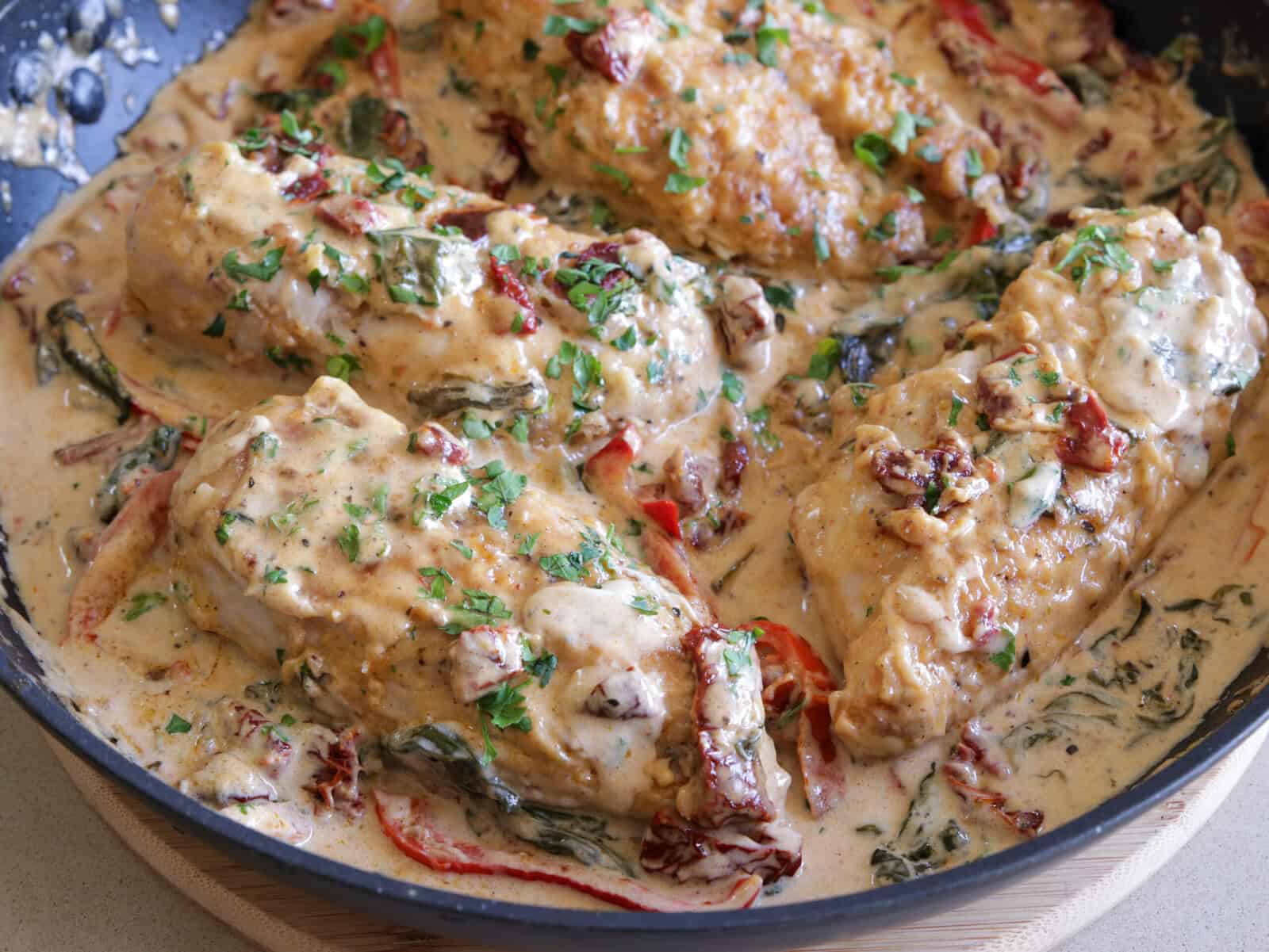 Chicken breasts simmering in a creamy sauce with spinach, sun-dried tomatoes, and herbs, garnished with chopped parsley in a skillet.