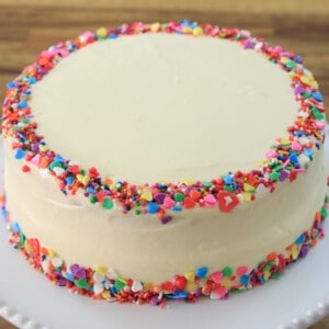 A round, frosted cake with smooth white icing, decorated with colorful sprinkles around the top and bottom edges, placed on a white plate with a wooden background.