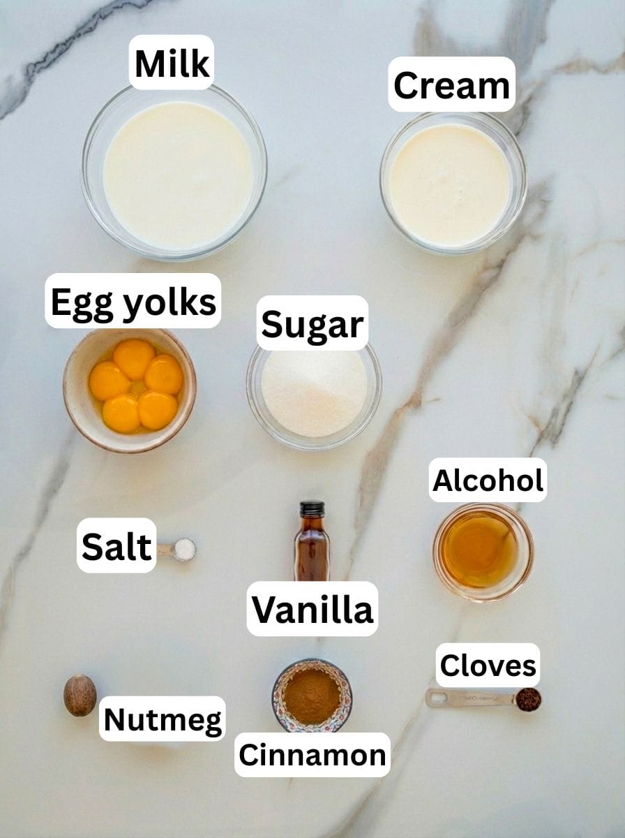 Overhead flat-lay of eggnog ingredients arranged on a white marble surface: bowls of milk, cream, sugar, and egg yolks, small dishes of cinnamon and cloves, a whole nutmeg, a bottle of vanilla extract, a pinch of salt, and a small glass of alcohol, each ingredient clearly labeled.