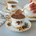 A hand holds a spoonful of thick hot chocolate above an ornate floral teacup filled with hot chocolate, with two more matching teacups and a striped napkin in the background.