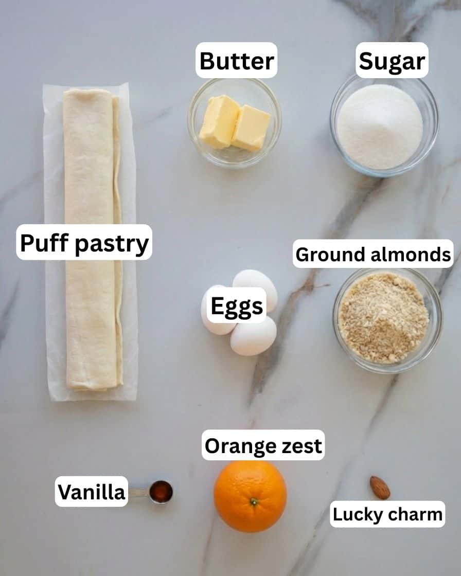 Eight baking ingredients are arranged on a marble surface: a roll of puff pastry, two cubes of butter in a bowl, a bowl of sugar, a bowl of ground almonds, three eggs, vanilla extract, an orange, and a whole almond.