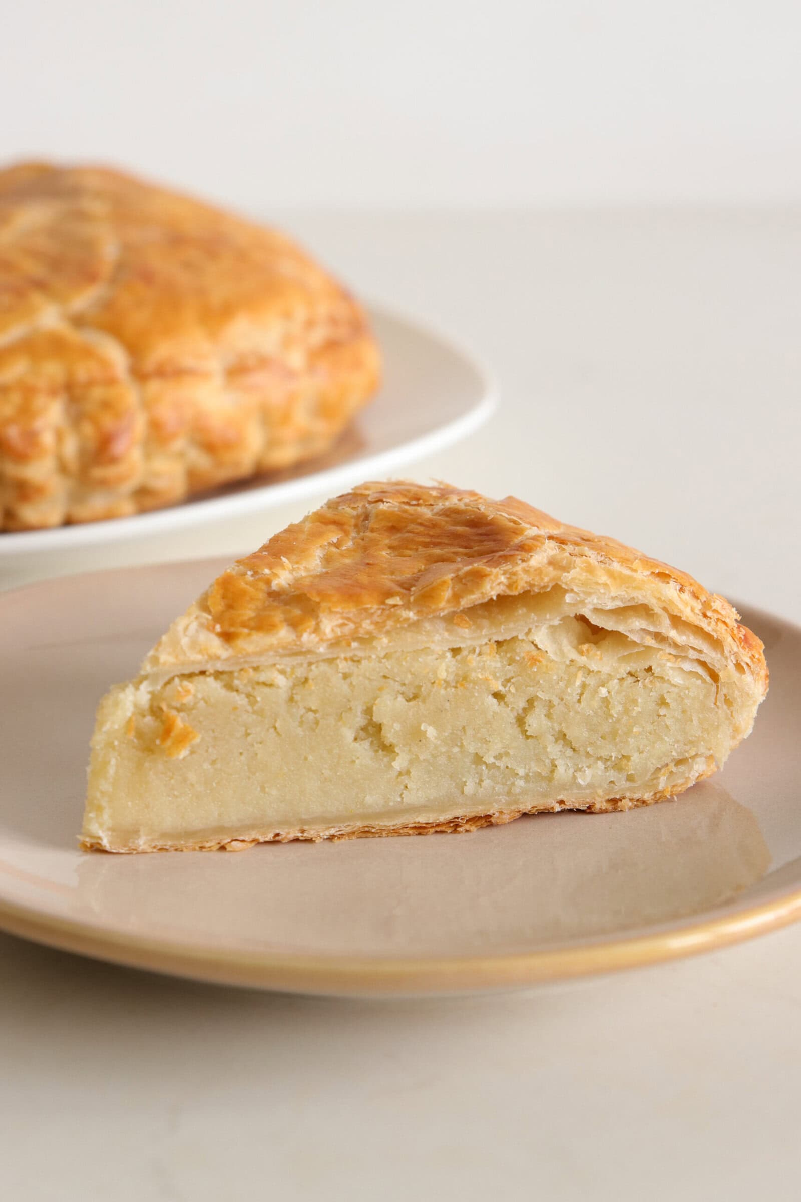 A slice of golden, flaky galette des rois with a creamy filling sits on a beige plate, with the rest of the round pastry visible in the background on a white plate.
