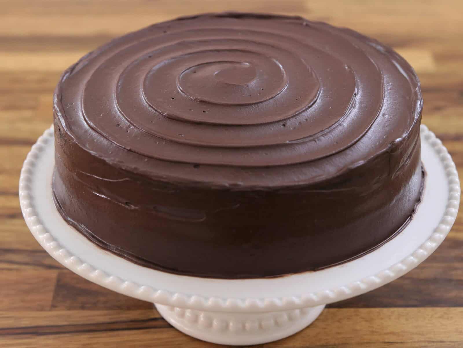 A round chocolate cake with smooth, glossy chocolate frosting and a spiral pattern on top, displayed on a white cake stand over a wooden surface.