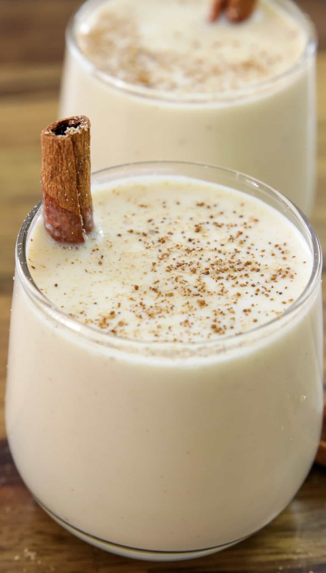 A glass of creamy eggnog topped with ground nutmeg and garnished with a cinnamon stick sits on a wooden surface, with a second glass visible in the background.