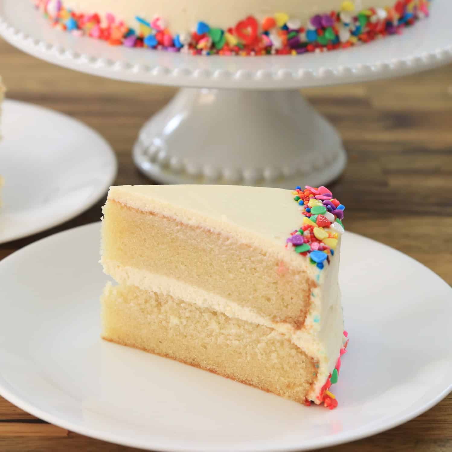 A slice of two-layer vanilla cake with white frosting and colorful sprinkles on the edge, served on a white plate. The whole cake, also decorated with sprinkles, is in the background on a cake stand.