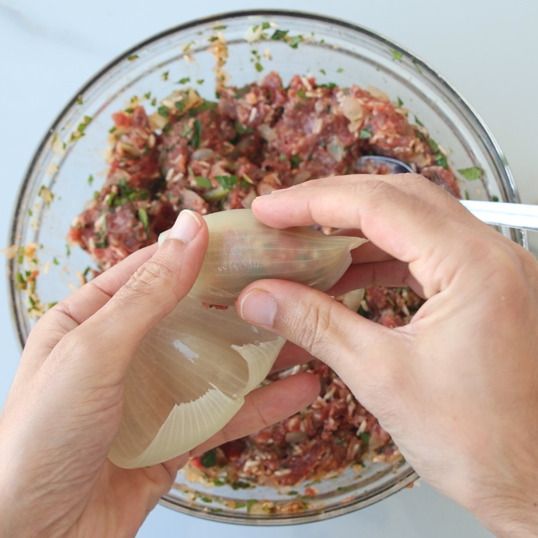 Hands separating layers of a large onion in front of a bowl filled with ground meat, herbs, and vegetables, preparing to fill the onion with the meat mixture.