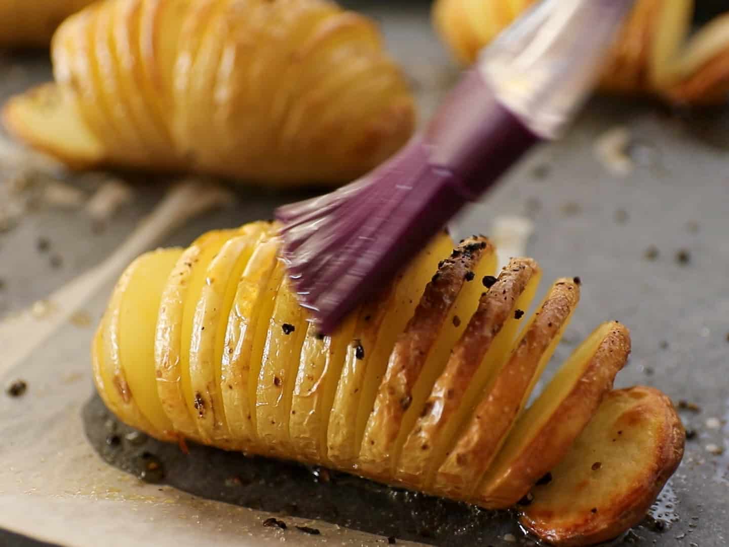 A close-up of a sliced, baked potato being brushed with oil or butter, sprinkled with black pepper, on a baking sheet.