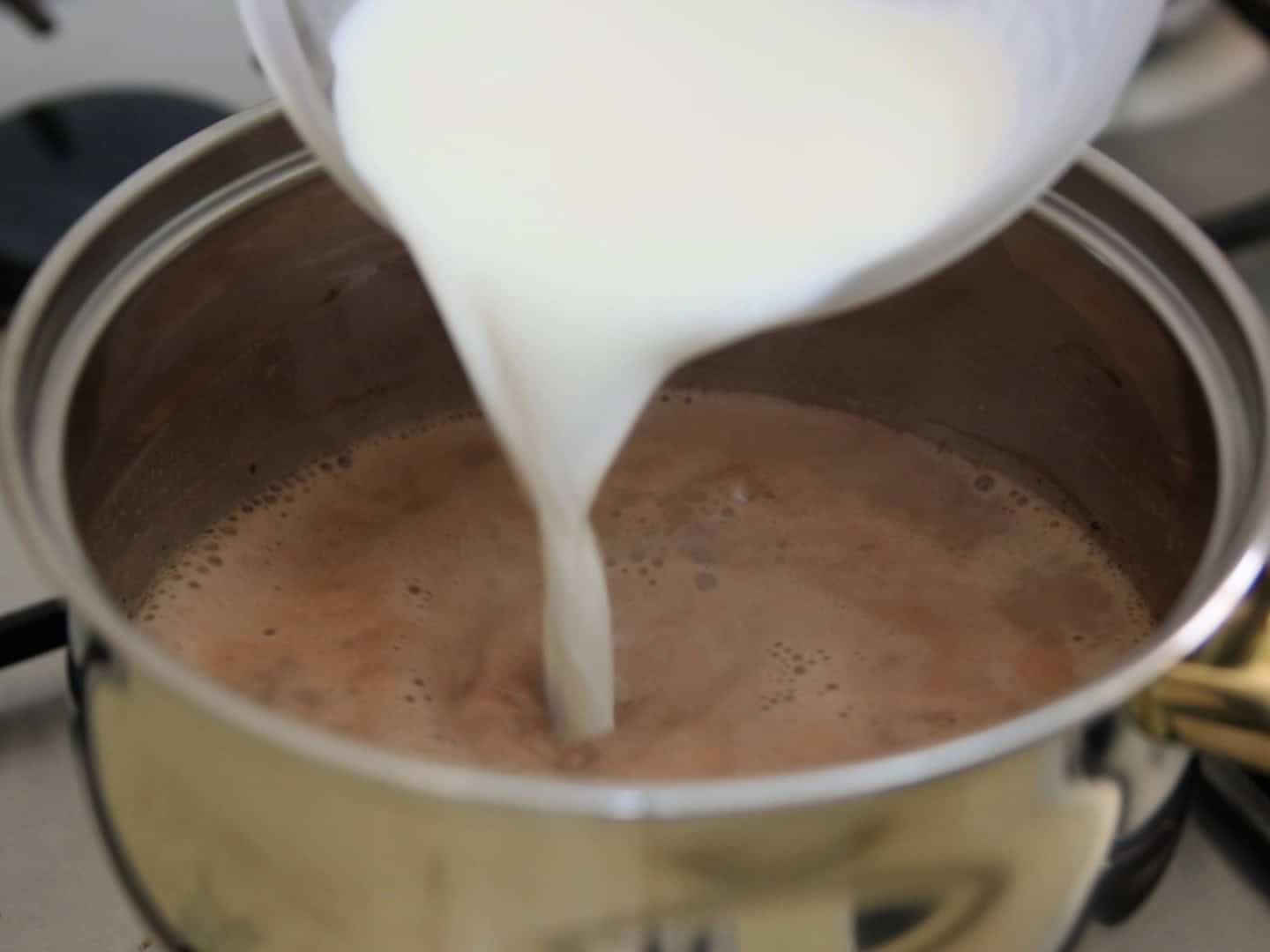 A close-up of milk being poured from a container into a saucepan of hot chocolate on a stovetop.