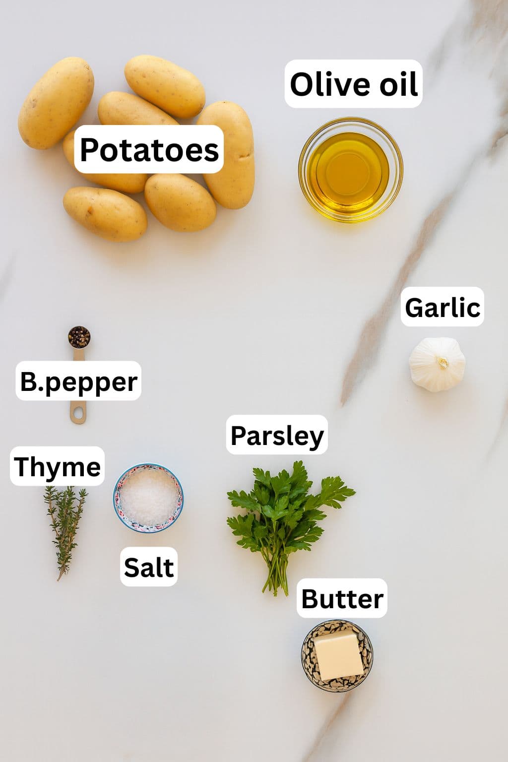 A marble surface with labeled ingredients: whole potatoes, a bowl of olive oil, a garlic bulb, fresh parsley, a pat of butter, salt, black pepper, and sprigs of thyme.