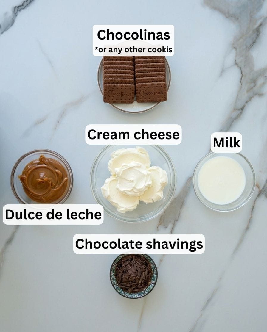 Five labeled bowls and plates with ingredients: a stack of Chocolinas cookies, a bowl of cream cheese, a bowl of milk, a bowl of dulce de leche, and a small bowl of chocolate shavings on a light surface.