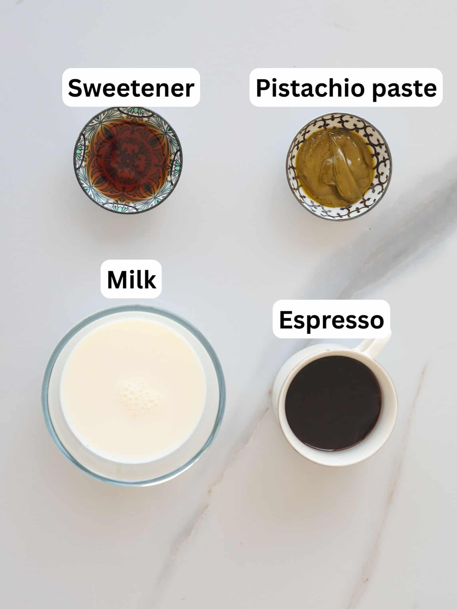 Four labeled ingredients on a light surface: a bowl of sweetener, a bowl of pistachio paste, a glass of milk, and a cup of espresso.