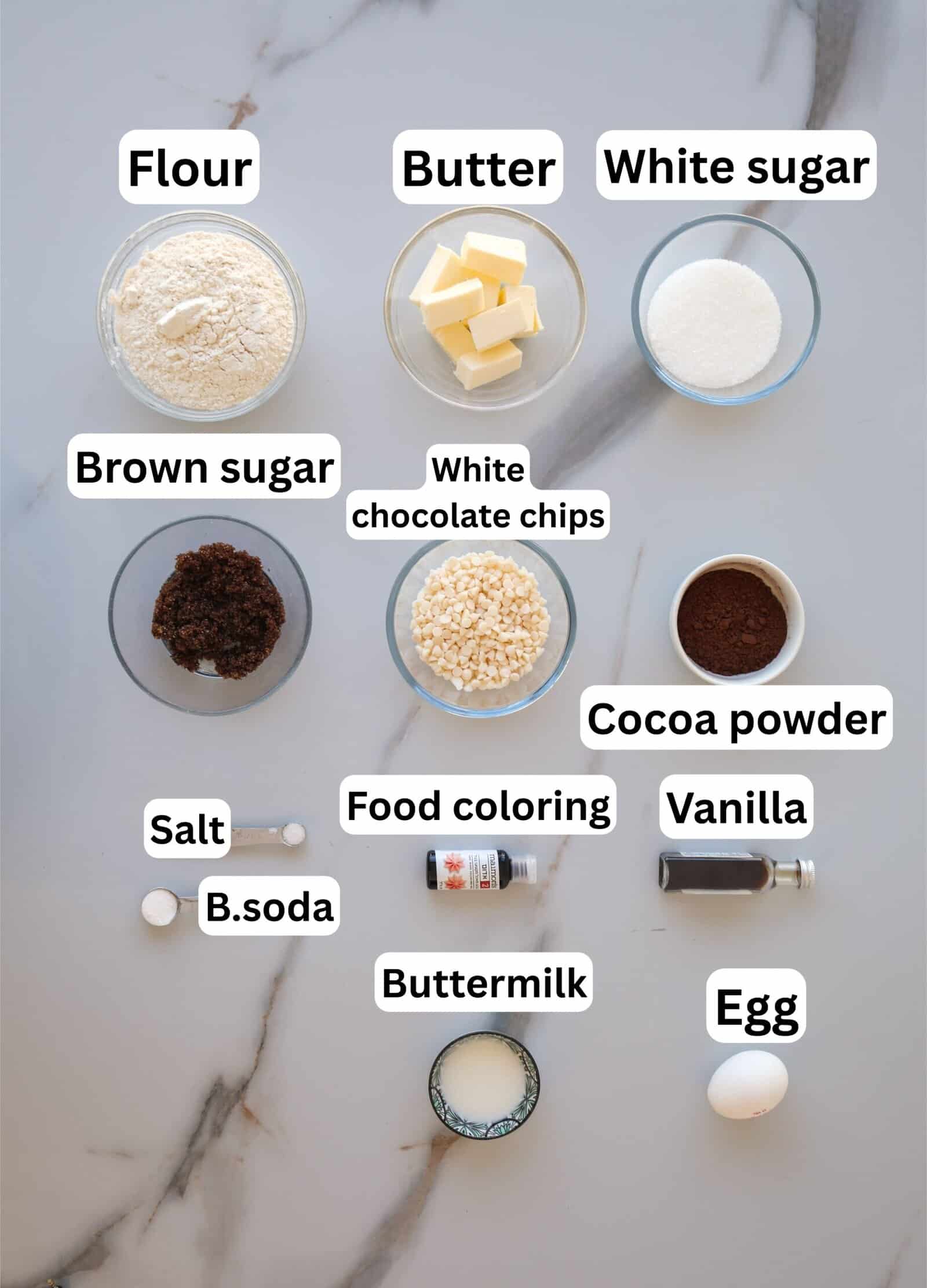 Baking ingredients arranged on a marble surface, each labeled: flour, butter, white sugar, brown sugar, white chocolate chips, cocoa powder, salt, baking soda, food coloring, vanilla, buttermilk, and an egg.