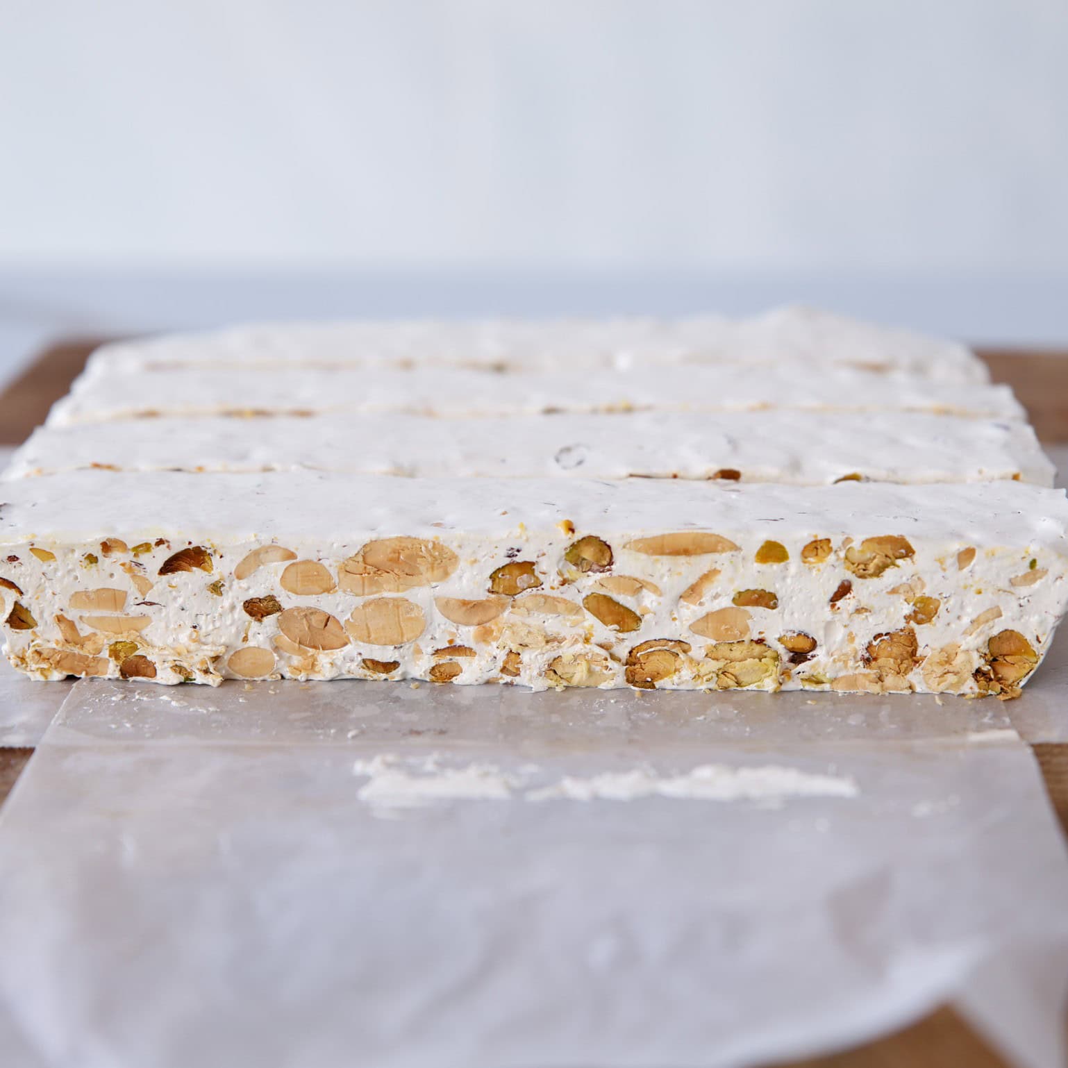A close-up of nougat (Italain torrone) with visible whole nuts, sliced into rectangular pieces and resting on a sheet of wax paper. The nougat is white and chewy, with a smooth surface and a dense nut filling.