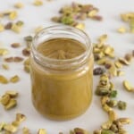 A glass jar filled with creamy pistachio butter sits on a white surface, surrounded by scattered whole pistachio nuts.